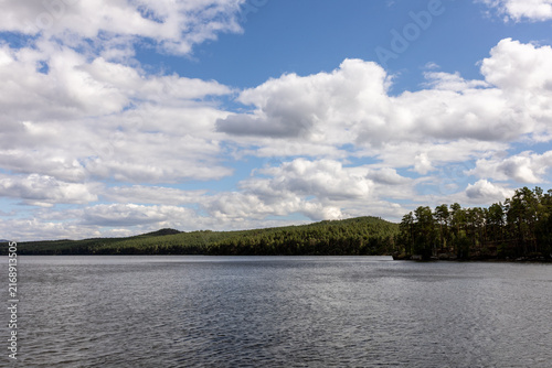 Scenic lake landscape with calm rippling water, lush green shoreline, resort buildings and gazebos surrounded by trees, with mountains visible in the distance under a dramatic cloudy sky. Peaceful out