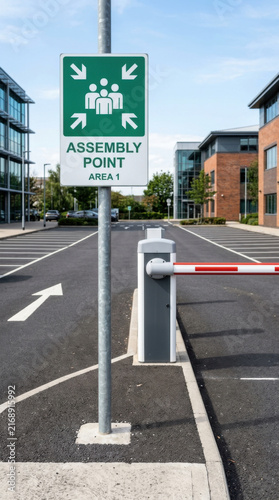 Assembly Point Sign In Front Of Modern Office Buildings And Barrier