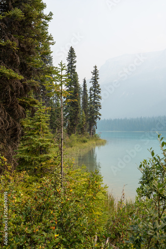 Emerald Lake, Canada - August 14, 2026: Emerald lake surrounded by forest and mountains under hazy sky caused by wildfire smoke. Canoes on the calm water create an atmospheric and moody summer scene.