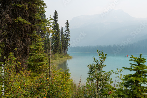 Emerald Lake, Canada - August 14, 2026: Emerald lake surrounded by forest and mountains under hazy sky caused by wildfire smoke. Canoes on the calm water create an atmospheric and moody summer scene.