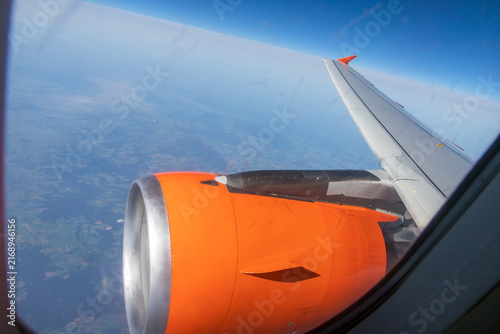 An aerial view from passenger aircraft window showing  bright orange engine cowl against a landscape of patchwork fields below. Looking down from a cruising altitude through passenger cabin window. 
