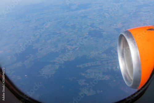 An aerial view from passenger aircraft window showing  bright orange engine cowl against a landscape of patchwork fields below. Looking down from a cruising altitude through passenger cabin window.
