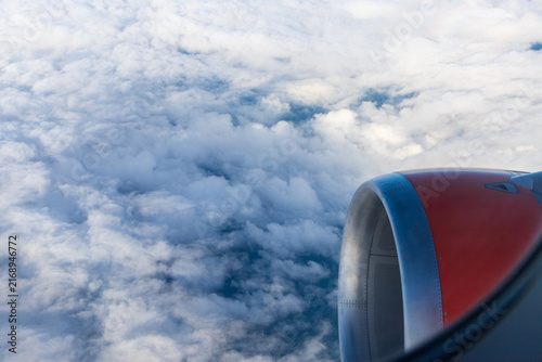 A view from an airplane window looking down past a metallic and red engine nacelle at a dense layer of fluffy white clouds. Vantage point, a commercial flight. An aerial photograph from high altitude.