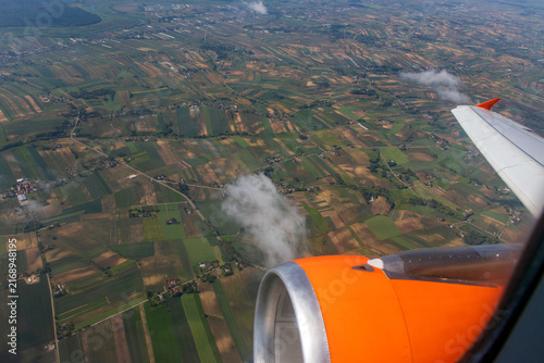 On the airplane an aircraft engine is visible from a window seat, looking down at an aerial view of green fields and patchwork farmlands. Patchwork agricultural landscape and scattered clouds below. 