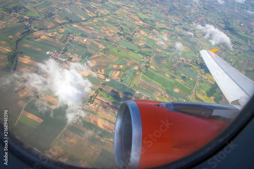 
On the airplane an aircraft engine is visible from a window seat, looking down at an aerial view of green fields and patchwork farmlands. Patchwork agricultural landscape and scattered clouds below. 
