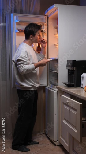 Vertical video young man opening the refrigerator door, looking inside for food and eating a snack during a late night craving in the kitchen