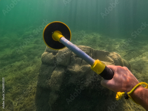 Quadro su tela First person view of diver hand holding handheld underwater metal detector searching for lost treasure near submerged stone structure in clear green lake water