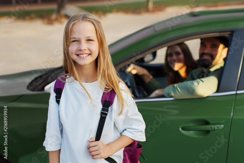 Obraz na plátne Happy schoolgirl with backpack standing in front of family car and smiling at ca