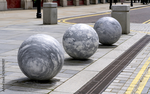 Three large marble balls sit on urban street pavement. Grey granite spheres form decorative street furniture in modern city district. Minimalist stone bollards line sidewalk near drainage grate.