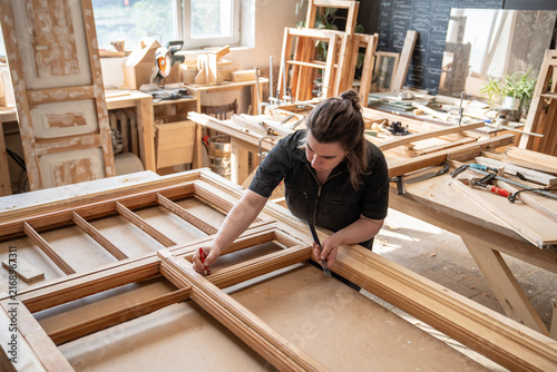 Fotografie Female craftswoman emerged in work on restoring antique window frame at workbench in workshop, talented woman carpenter measures wooden frame parts and making marks on wood planks with pencil