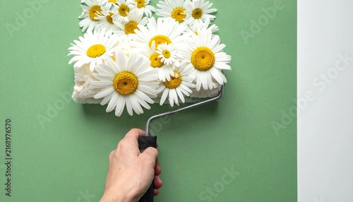 Hand holding paint roller covered in white daisy flowers leaving floral stroke on light green background