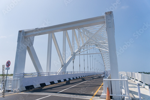 White steel arch bridge spans calm waters under clear blue sky. Road stretches forward with smooth asphalt and defined lane markings. Pedestrians' shadows cast long across the bridge's clean surface
