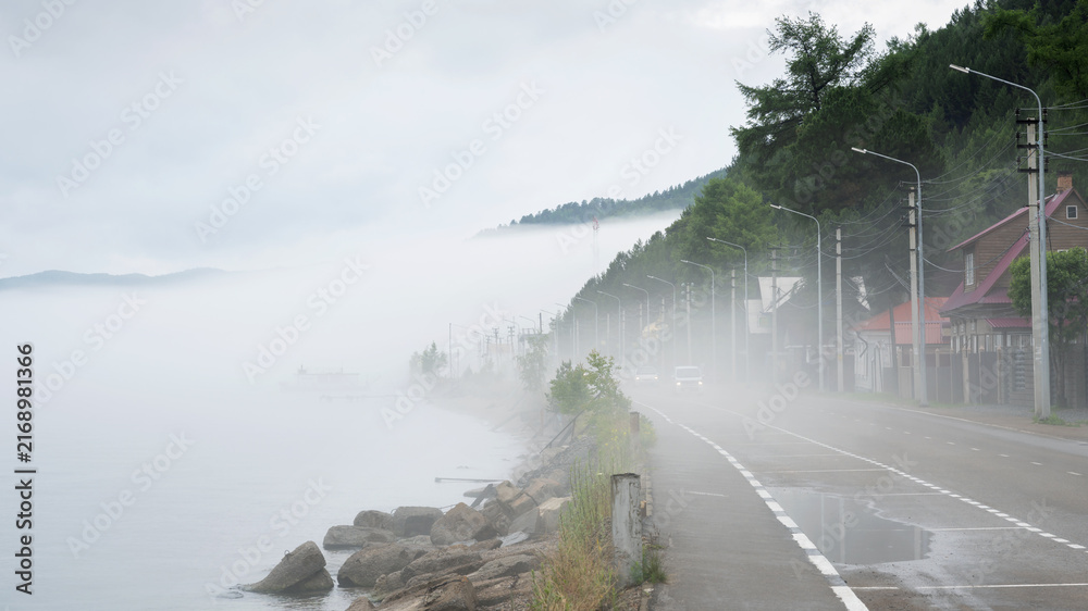 Fototapeta premium Thick fog over a coastal road with driving cars at Lake Baikal, Russia. Low visibility weather conditions on a lakeside highway with houses and streetlights.