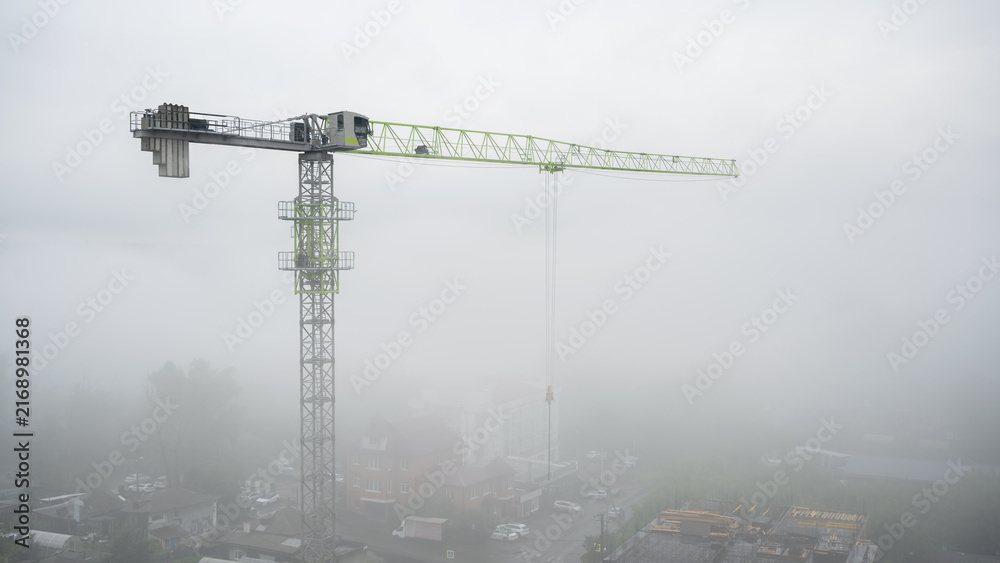 Naklejka premium Tower crane at a construction site covered in heavy fog. Urban building development in misty weather. Copy space for text