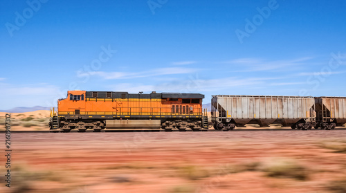 Freight Train in the Desert: An orange locomotive pulls a line of freight cars across a vast, arid desert landscape under a clear blue sky.