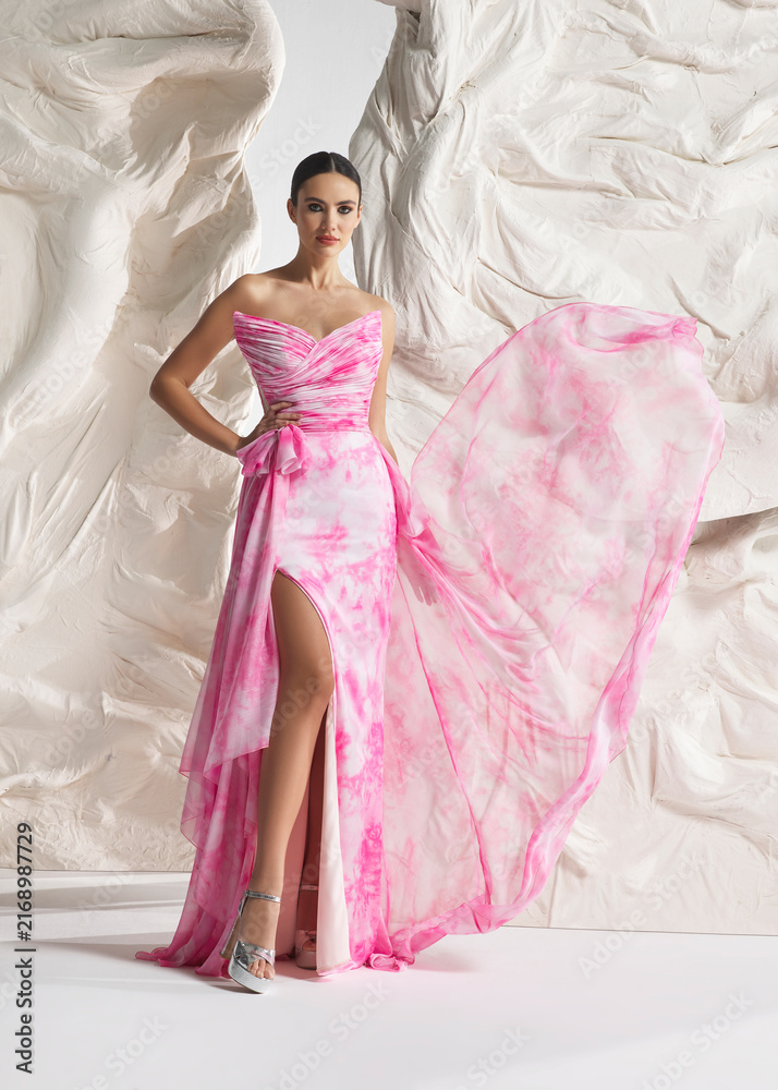 Fototapeta premium Full length shot of elegant woman wearing luxury pink floral watercolor floral organza gown with high-low train and tiered frills posing gracefully against sculptural white studio wall
