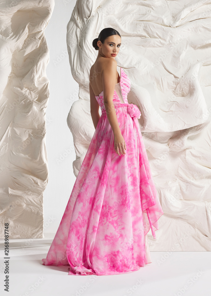 Fototapeta premium Full length shot of elegant woman wearing luxury pink floral flowing watercolor floral gala gown with ruffled train and corset posing gracefully against sculptural white studio wall backdrop