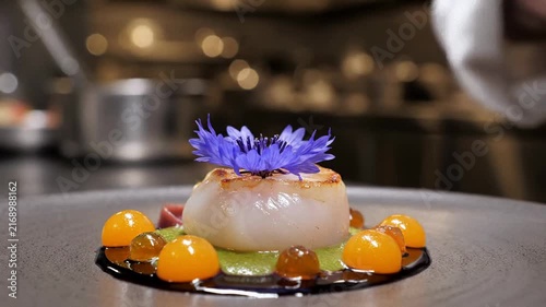 Professional chef using tweezers for plating, gourmet seared scallop with edible purple flowers, fine dining restaurant kitchen, luxury culinary art