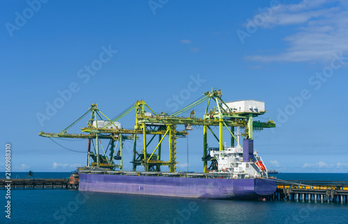 Large purple bulk carrier cargo ship docked at an ocean port terminal under big green crane structures, loading or unloading freight against a clear blue sky and calm water background