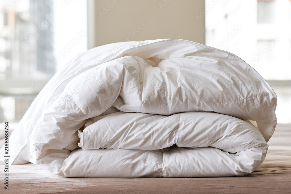 Naklejka premium White folded winter blanket stack with soft quilted layers on wooden surface, bright contemporary bedroom background, shallow focus emphasizing cozy textile texture and craftsmanship