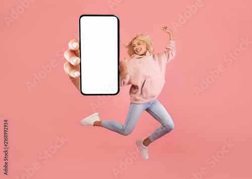 Happy Young Woman Jumping Excitedly With Smartphone Mockup In Hand, Standing On Pink Background And Showing Empty Screen. Cheerful Millennial Female Expressing Joy And Positivity In Studio.