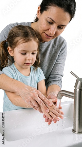 A mother teaches her daughter to wash her hands in a bathroom sink. She is gently guiding her daughter's hands under the running water