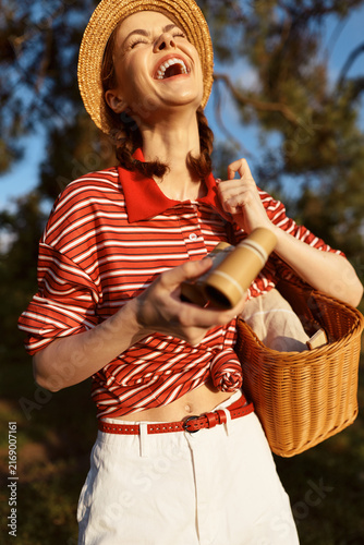 Konstfotografi A joyful young woman in a striped red shirt laughs loudly while carrying a wicker basket outdoors on a sunny day, capturing a carefree summer moment