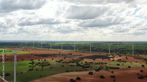 Large wind turbines stand tall against a backdrop of green fields and brown earth. The calm afternoon sun shines down, illuminating the landscape with vibrant colors.