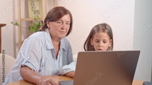 A grandmother and her granddaughter sit together at a table using a laptop. They look focused and engaged while exploring the screen. The home is bright and welcoming.