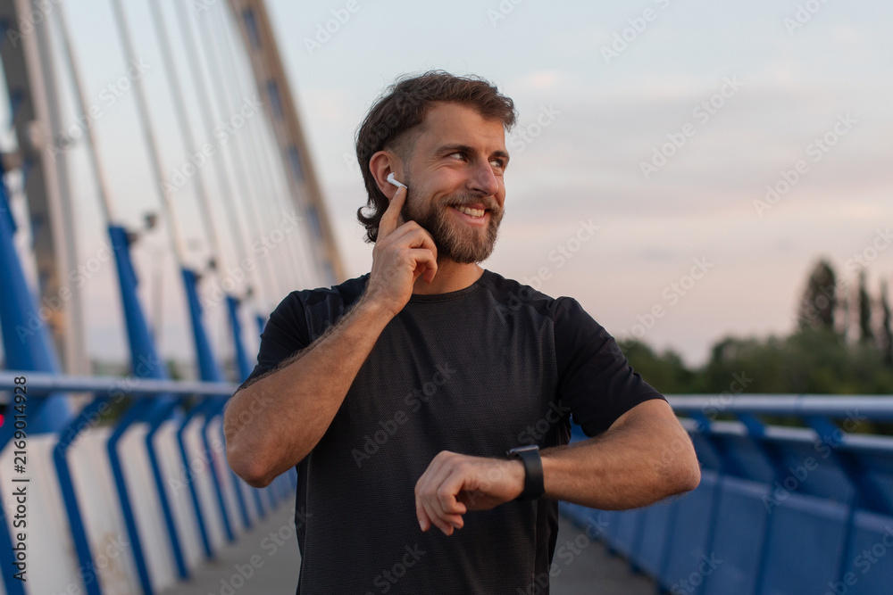 Naklejka premium Smiling sporty man pausing during his run to check time on his smartwatch while standing on modern bridge and touching earphones