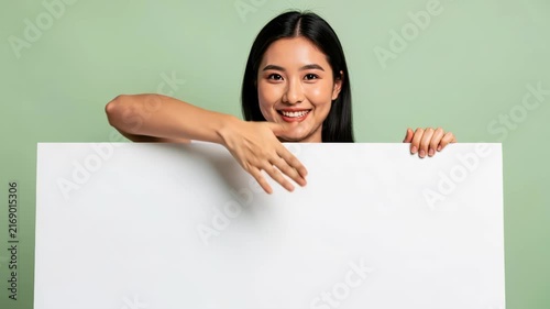 Asian Woman With Blank Sign: A beautiful young Asian woman smiles confidently as she holds a blank white sign, inviting custom messages or promotions, set against a soft green backdrop.