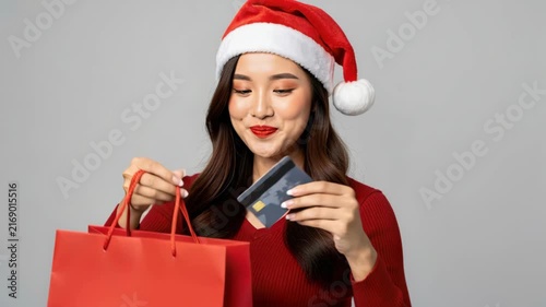 Christmas Shopper with Credit Card: A smiling woman wearing a Santa hat holds a credit card over a red shopping bag, ready for holiday purchases.