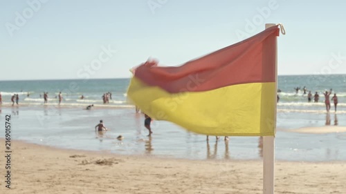 People enjoy a busy beach day under a clear sky. Waves crash on the shore while a flag flutters in the warm breeze, marking the coastline for beachgoers.