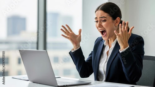 Frustrated Office Worker: A woman in business attire throws her hands up in despair and screams in frustration while looking at her laptop screen in an office setting.