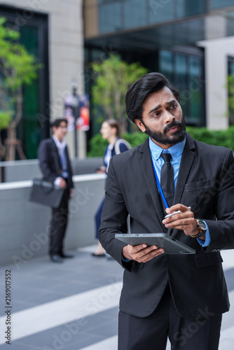 A bearded Indian businessman annotates a plan on the tablet with a digital pen outside a glass office tower in a modern Asian financial district, the shallow depth isolates him cleanly