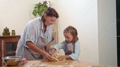 A woman and a girl are making dough together in a kitchen. They mix and knead the dough on a wooden table. Ingredients and tools are present on the table.