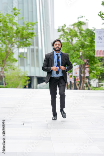 A young Indian businessman keeps running while glancing at his wristwatch along a sunlit walkway between contemporary corporate towers, clipped hedges streak past on both sides