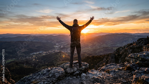 A person stands with arms raised on a mountain peak at sunset, celebrating a triumphant achievement, with breathtaking panoramic views of rolling hills below