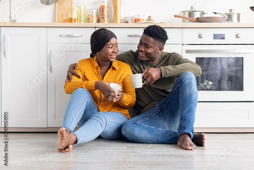 Quadro su tela Smiling Black Couple Sitting On Kitchen Floor, Holding Coffee Cups, Enjoying Quality Time Together, Surrounded By Cozy Kitchen Setting