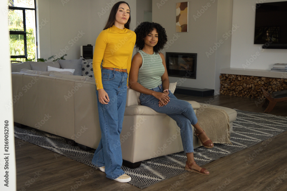 Fototapeta premium Diverse female friends posing in living room by beige sectional sofa, wearing jeans and tops