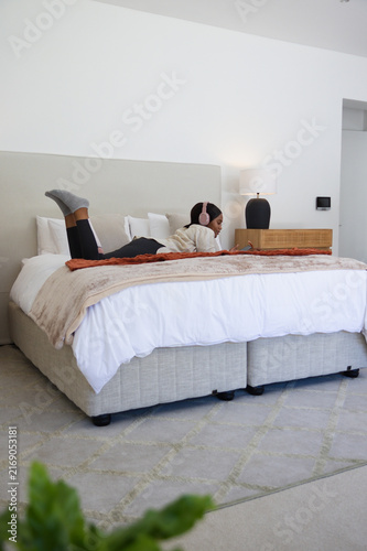 Woman lying on king-size bed using smartphone with pink headphones in minimalist bedroom