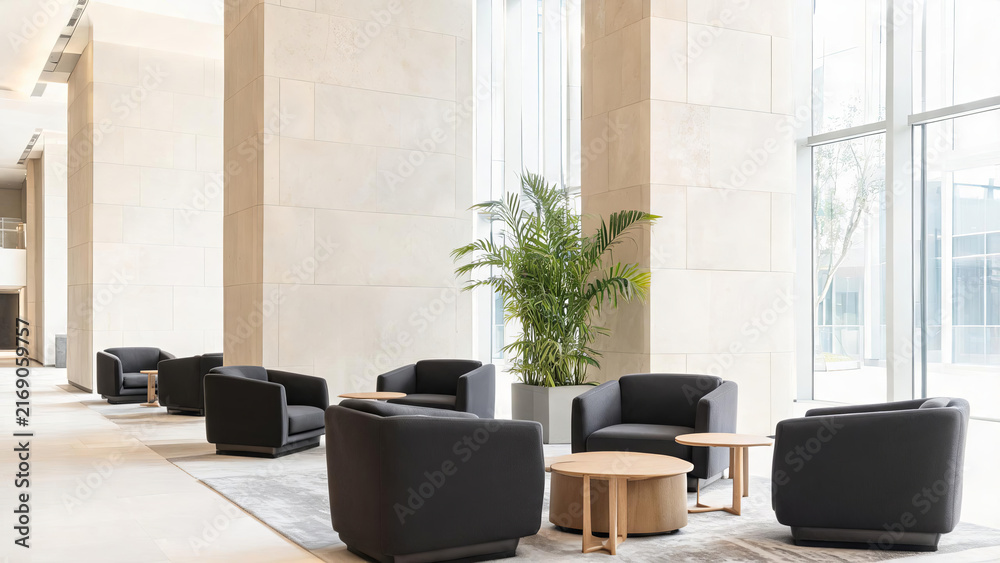 Fototapeta premium A bright modern indoor lobby seating area with dark armchairs, small tables, a large potted plant, and light stone walls and floors.
