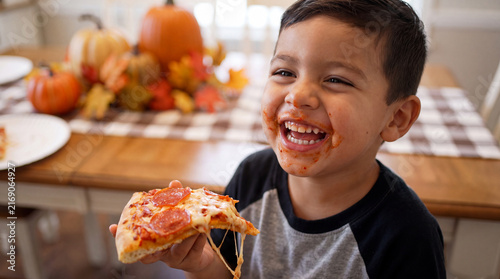 A joyful Latino toddler boy laughing while eating a cheesy pepperoni pizza slice at a National Pizza Month kitchen party, ideal for marketing and social media.
