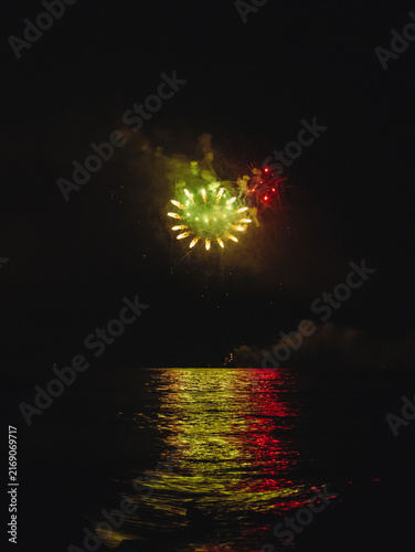 Canvas Print Bright green and red pyrotechnic fireworks burst, illuminating the thick smoke clouds and reflecting vibrant light on the dark ocean waves