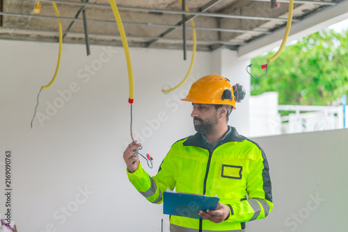 Electrician inspecting electrical wiring on ceiling with clipboard at house construction site