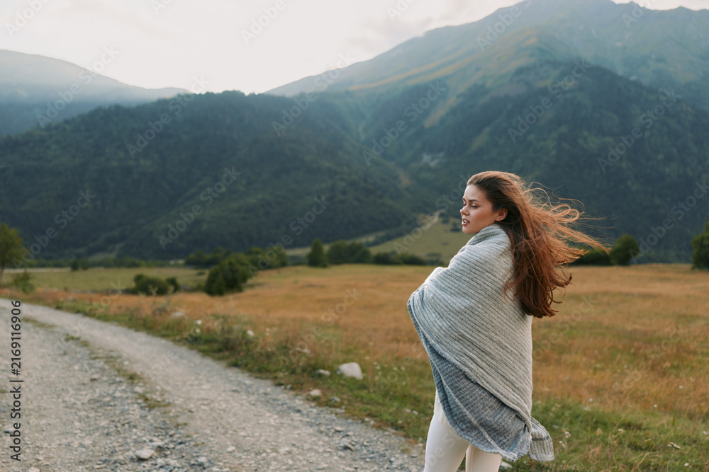 Fototapeta premium Woman standing on a dirt road in a valley wrapped in a light shawl looking to the side with wind in her hair Mountain view outdoor scene travel lifestyle calm mood
