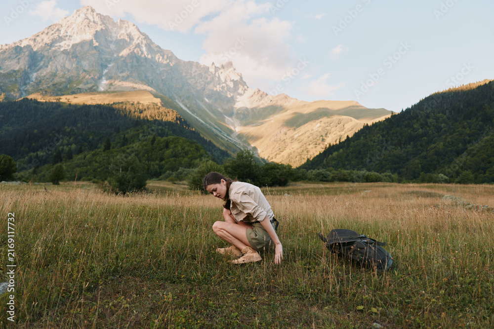 Naklejka premium Woman in outdoor meadow with distant mountains, crouching amid grass, backpack nearby, embracing sunlight and travel vibe, natural portrait of exploration