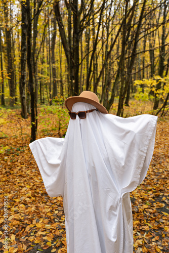 A playful ghost figure draped in a white sheet, wearing a tan hat and dark sunglasses, standing among trees covered with yellow fall leaves in a wooded park