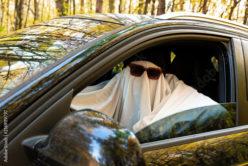 Person wearing ghost costume driving car while holding paper map, searching for direction and navigating route. Sunglasses cap and White fabric disguise covers the body and face, creating humorous