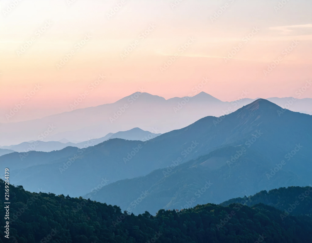 Fototapeta premium Mountain landscape at sunrise with layered blue ridges, forested hills, and pastel pink sky creating peaceful wilderness scene for nature travel imagery and outdoor background use